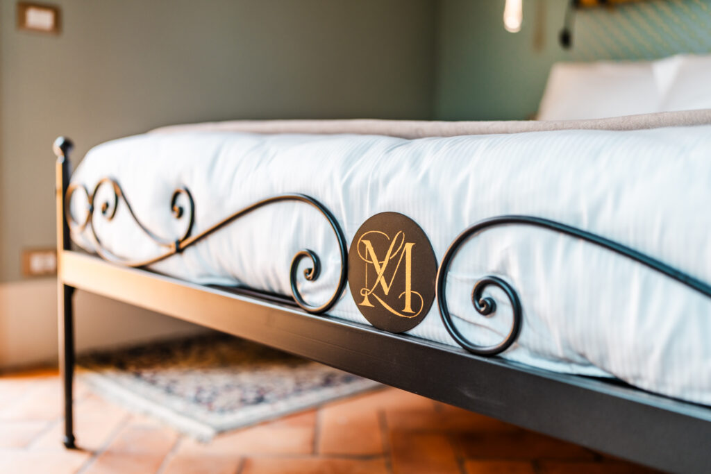 Detail of wrought-iron bed with Villa MonteLandi monogram, terracotta floor, and soft linens — symbol of bespoke Tuscan design and refined hospitality.