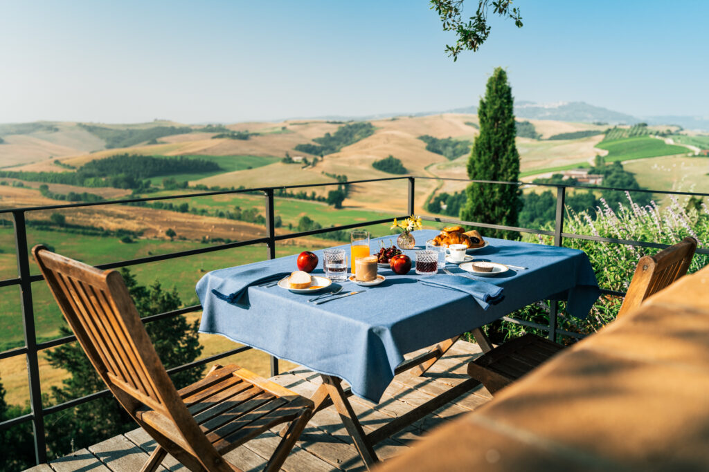 Elegant breakfast setup on a panoramic terrace overlooking the Tuscan hills and Montalcino, surrounded by cypress trees and lush greenery