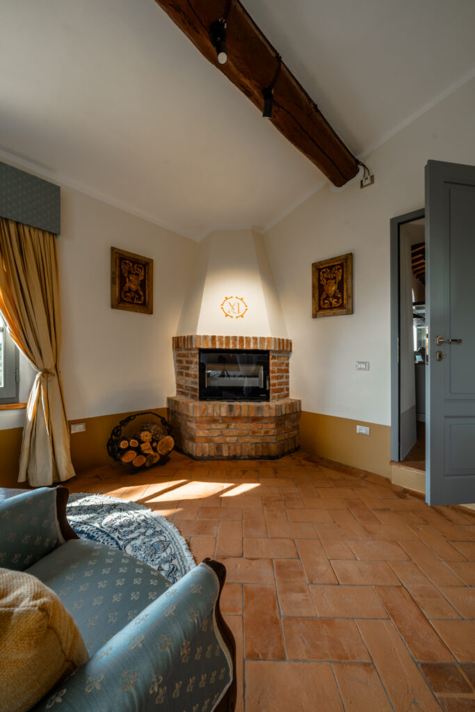 Cozy corner with a brick fireplace in the Vento di Lavanda apartment at Villa Montelandi, featuring warm terracotta floors, wooden beams, and elegant Tuscan decor.