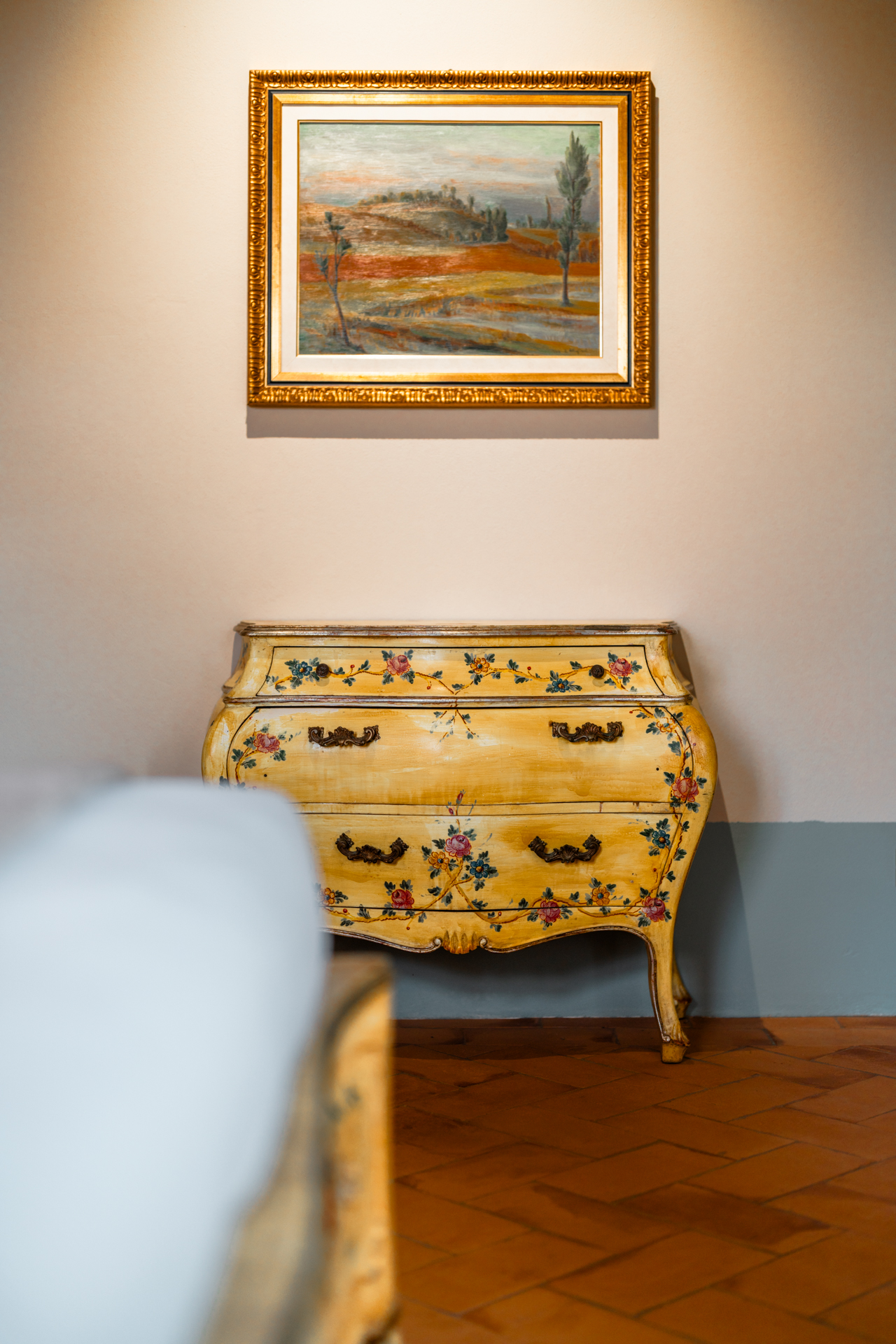 Detail from the Tramonto Toscano apartment at Villa Montelandi, featuring a hand-painted antique chest of drawers and a framed landscape painting inspired by the Tuscan countryside.