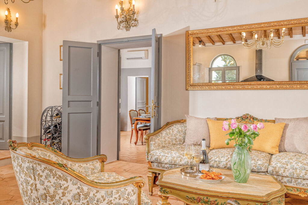 Elegant living room of the Tramonto Toscano apartment at Villa Montelandi, featuring antique floral furniture, golden mirror, and a cozy Tuscan atmosphere with wine and flowers on the table.