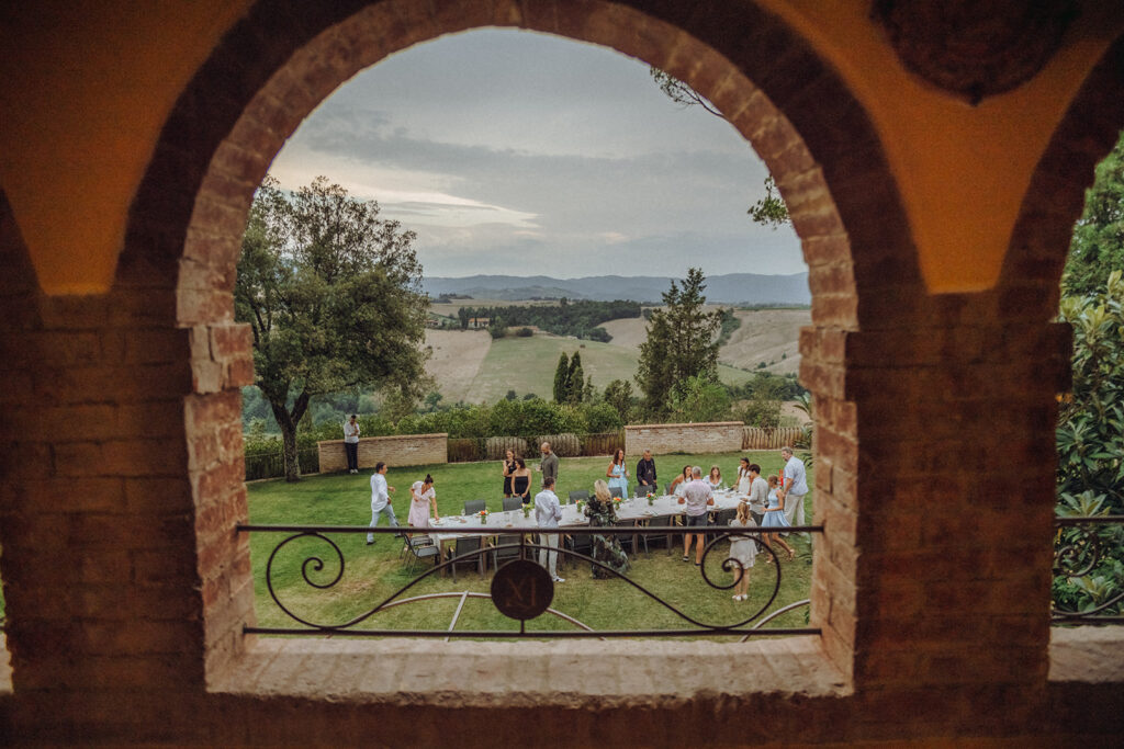 View through the arches of Villa Montelandi overlooking a garden dinner party with guests enjoying an evening gathering against the rolling hills of Tuscany.