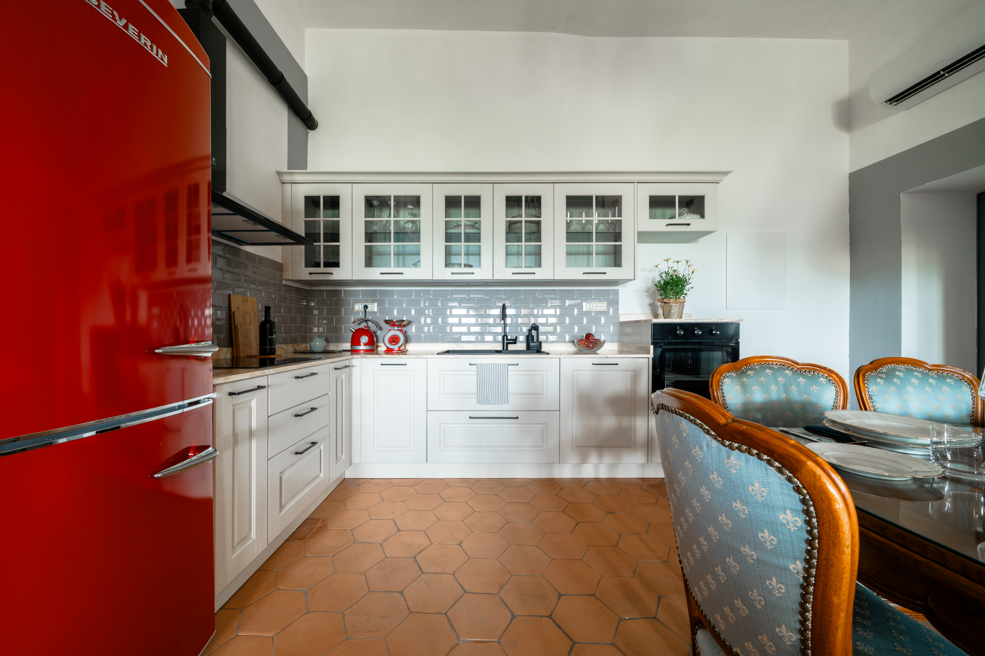 Elegant Tuscan kitchen at Villa MonteLandi with white cabinetry, red Smeg fridge, terracotta floor, and dining table set for guests — a refined blend of tradition and modern comfort in Tuscany.