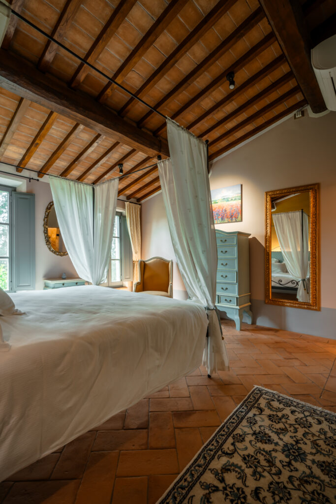 Cozy bedroom at Villa MonteLandi in Tuscany with canopy bed, terracotta floor, and wooden beam ceiling — a perfect blend of rustic charm and refined comfort.