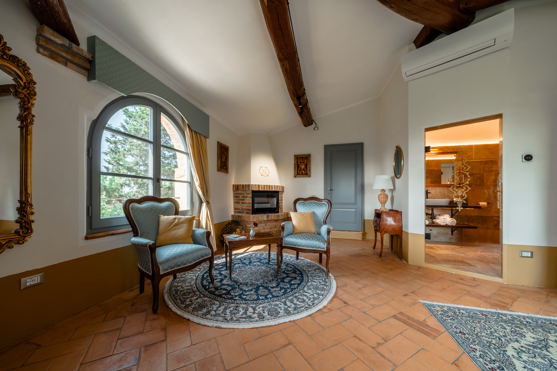 Elegant living area at Villa MonteLandi in Tuscany with arched window, antique armchairs, and brick fireplace — a refined space blending Tuscan tradition and comfort.