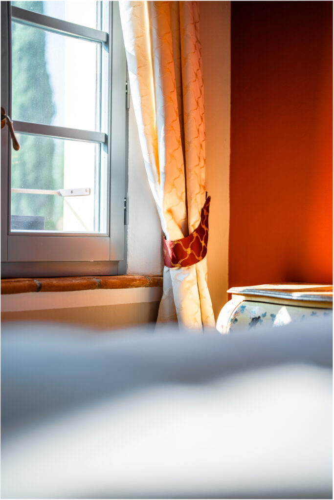 Detail of a sunlit window with elegant curtains and hand-painted furniture in a Tuscan bedroom at Villa MonteLandi — refined Italian craftsmanship and warmth.