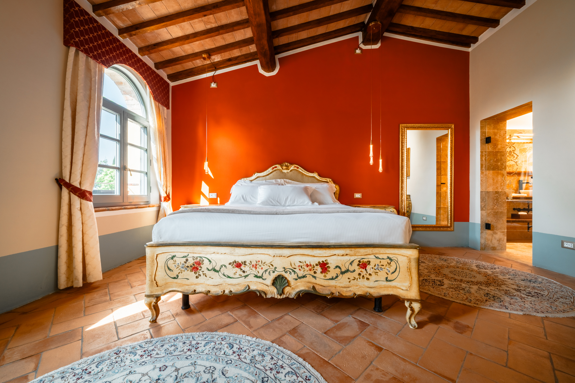 Elegant bedroom at Villa MonteLandi in Tuscany featuring an antique hand-painted bed, terracotta floor, and warm red accent wall — authentic Tuscan design with refined comfort.