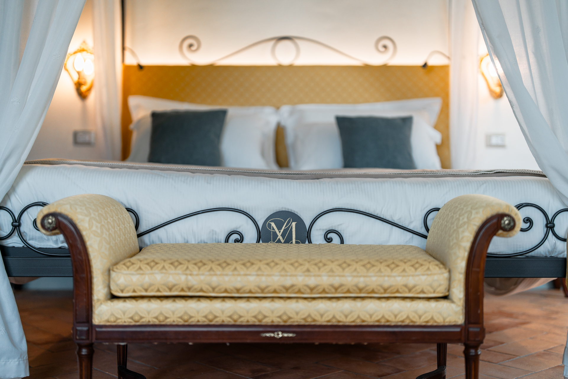 Elegant bedroom at Villa MonteLandi in Tuscany with wrought-iron bed, soft lighting, and upholstered bench, showcasing refined Italian craftsmanship and timeless comfort.