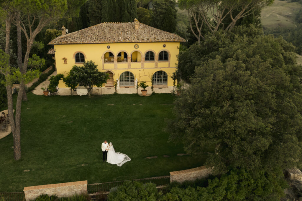 Aerial view of Villa Montelandi, a yellow Tuscan villa surrounded by gardens, with a bride and groom standing on the lawn.