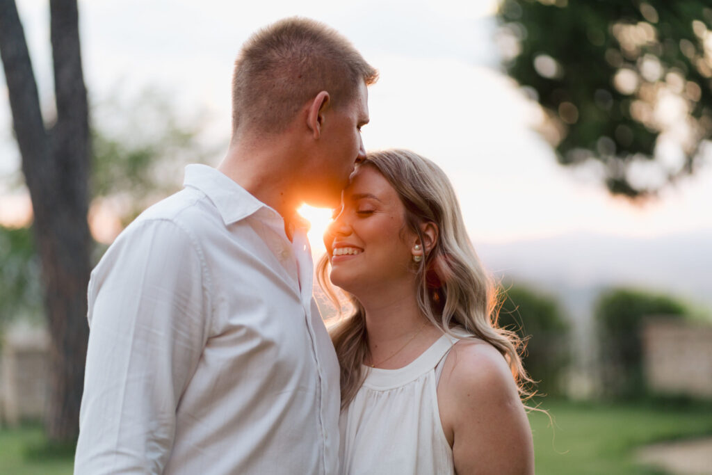 Couple sharing a tender kiss on the forehead at sunset in the gardens of Villa Montelandi, with warm golden light creating a romantic atmosphere.